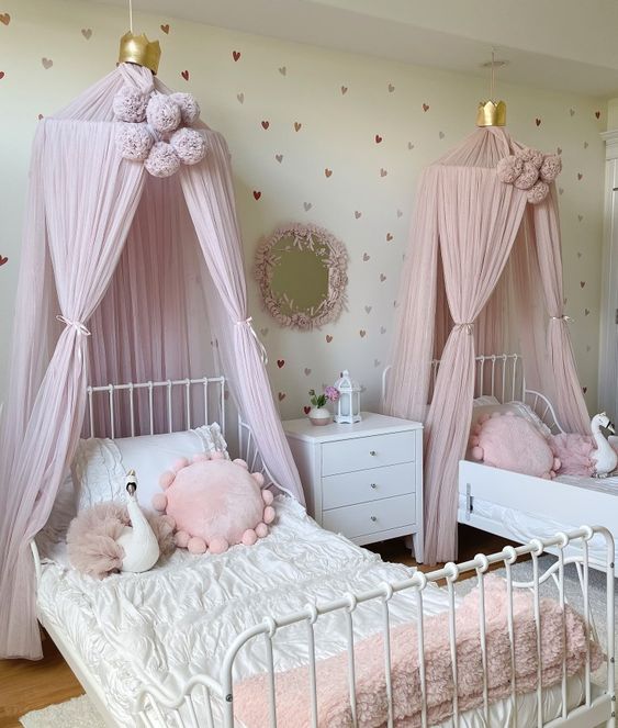 a princess style shared girls' bedroom with a white metal bed, pink canopies, pink pillows, a white nightstand and a mirror in a pink frame a princess-style shared girls' bedroom with a white metal bed, pink canopies, pink pillows, a white nightstand and a mirror in a pink frame