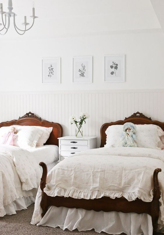 a refined French cottage shared girls' bedroom with paneling, dark stained carved wooden beds, a white nightstand and a gallery wall with botanicals