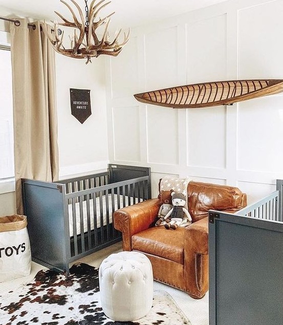 a shared farmhouse nursery with grey cribs, a leather chair, an antler chandelier and an animal skin rug