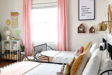 a stylish and catchy shared girls’ bedroom with black metal beds, white nightstands and dressers, pink curtains, macrame and house shelves