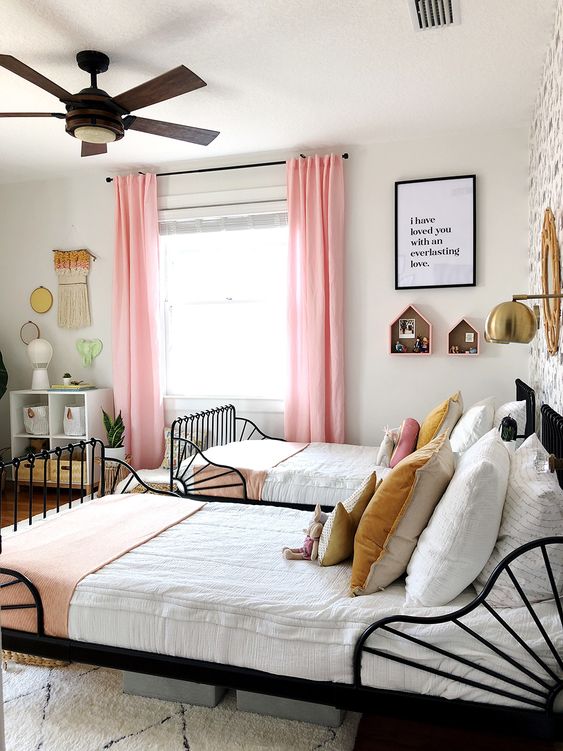 a stylish and catchy shared girls' bedroom with black metal beds, white nightstands and dressers, pink curtains, macrame and house shelves