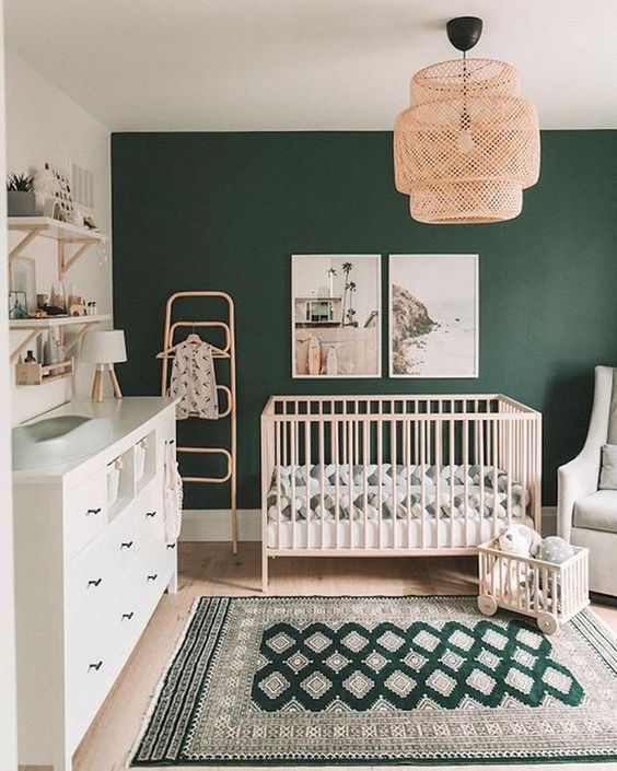 a stylish boho modern nursery with a hunter green accent wall, light-colored furniture, a woven chandelier and printed textiles