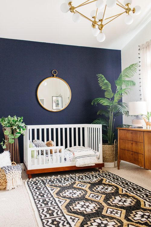 a stylish boho nursery with a navy accent wall, a white crib and a wooden sideboard, a printed rug and a round mirror plus statement plants