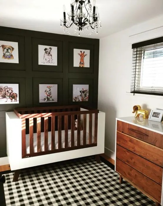 a stylish mid century modern nursery with a black accent wall with a gallery wall, a white crib, a stained dresser, a buffalo check rug and a black chandelier