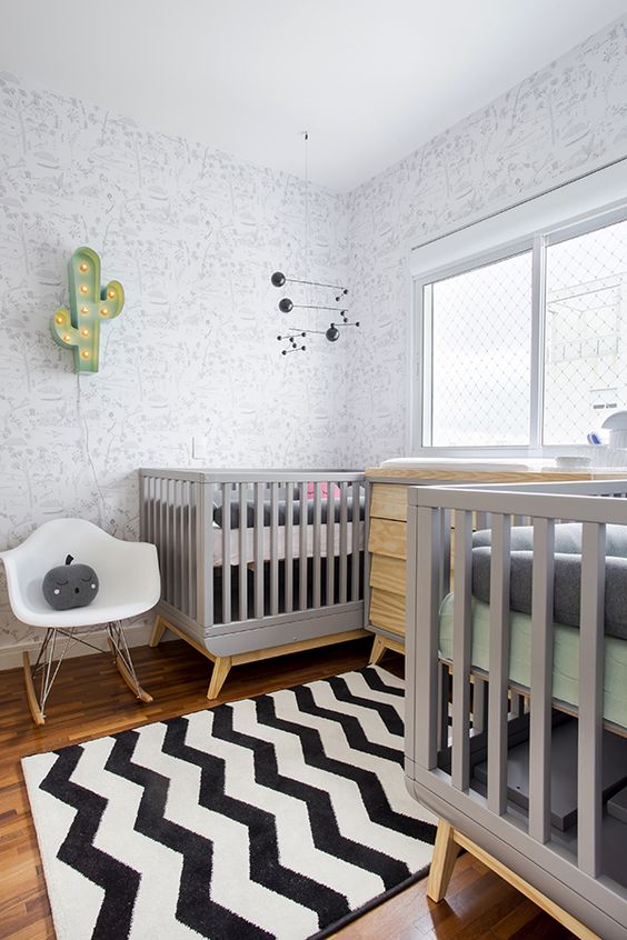 a stylish shared nursery with pinted wallpaper, grey and stained furniture, a printed rug and grey bedding