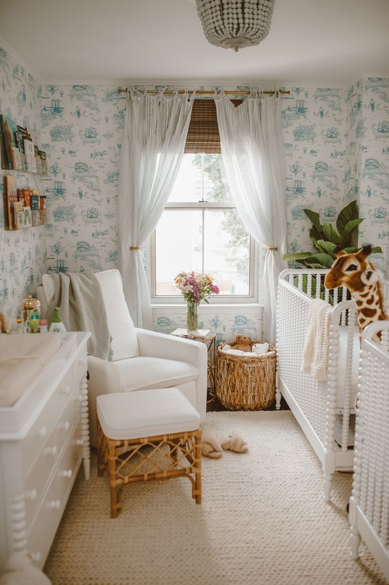 a tropical nursery with blue printed wallpaper, white furniture, a rattan stool, a beaded chandelier and a statement plant