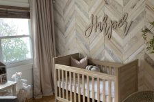 a warm neutral farmhouse nursery with a reclaimed wooden wall with a herringbone pattern, a bead chandelier, a cool rug and a stained crib