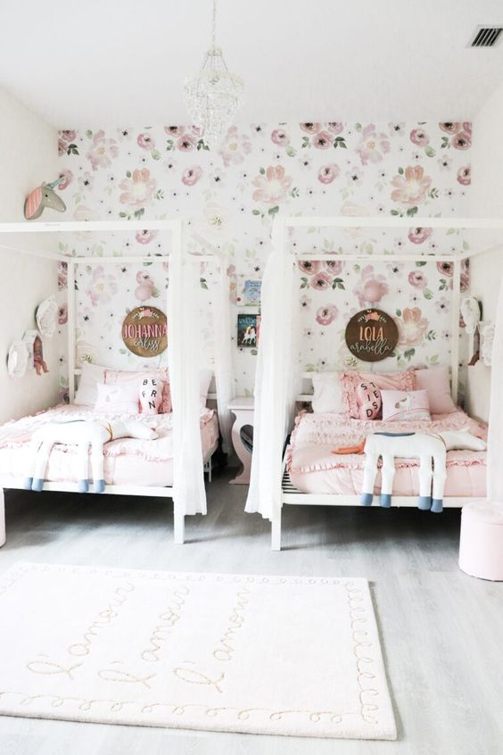 a whimsy shared girls' bedroom with a floral wallpaper wall, white beds with canopies, a nightstand, pink bedding, a rug and a pouf is wow