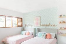 an adorable modern girls’ bedroom with a mint printed accent wall, grey upholstered beds, pink and pastel bedding, jute rugs, wooden shelves and names on the wall