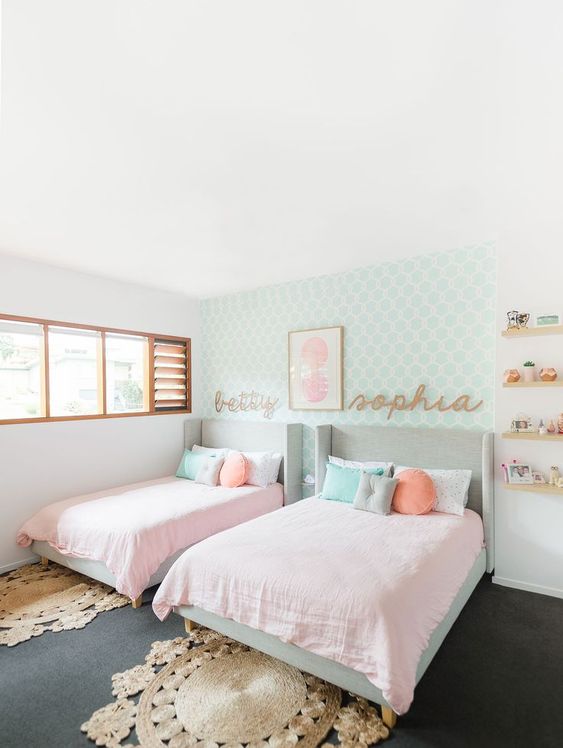 an adorable modern girls' bedroom with a mint printed accent wall, grey upholstered beds, pink and pastel bedding, jute rugs, wooden shelves and names on the wall