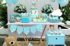 a blue dessert table with a backdrop done with clouds and blue balloons, a blue garland and blue and white floral arrangements