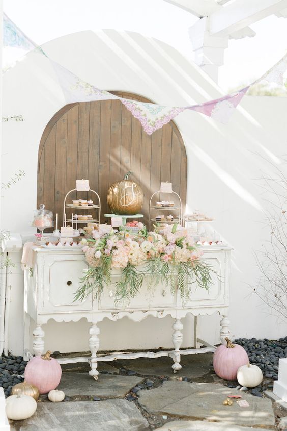 a delicate fall dessert table with a vintage sideboard, lots of painted pumpkins, a pink floral arrangement and a pink doily garland