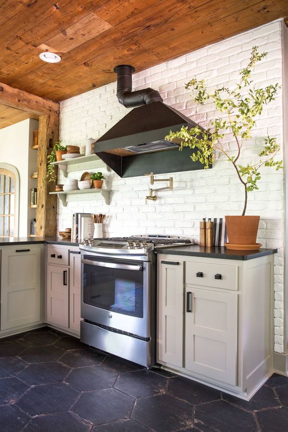 a dove grey kitchen with shaker style cabinets, black countertops, a white brick wall and potted plants plus a black hood