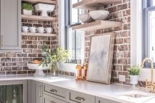 a dove grey kitchen with shaker style cabinets, white countertops, open shelves, red brick walls as a backsplash and brass touches