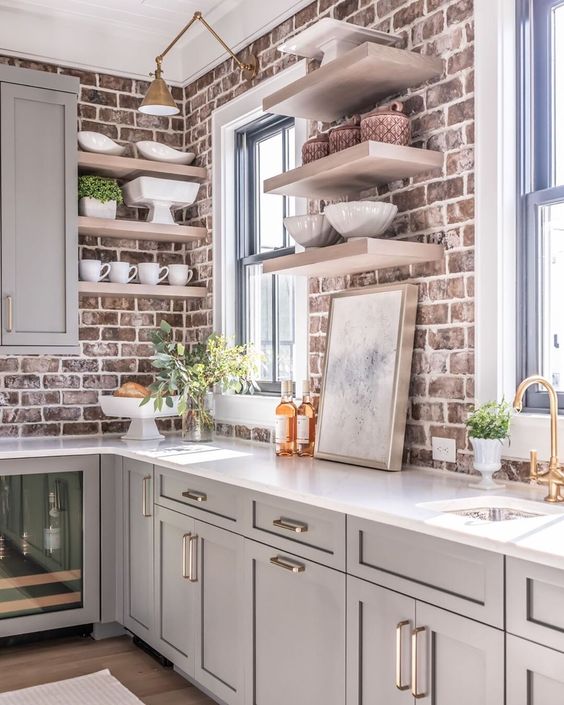 a dove grey kitchen with shaker style cabinets, white countertops, open shelves, red brick walls as a backsplash and brass touches