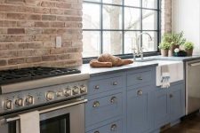 a dusty blue farmhouse kitchen with shaker style cabinets, grey stone countertops, a brick wall and a large window with much natural light
