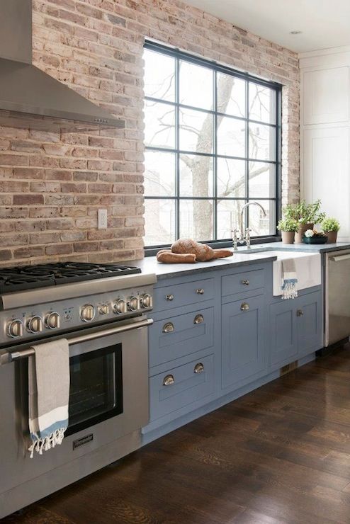 a dusty blue farmhouse kitchen with shaker style cabinets, grey stone countertops, a brick wall and a large window with much natural light
