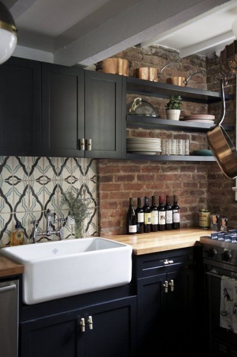 a moody kitchen with black cabinets, light-colored butcherblock countertops with a red brick backsplash