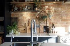 a navy kitchen with white stone countertops, open shelves, greenery and pendant lamps plus a red brick wall as a backsplash