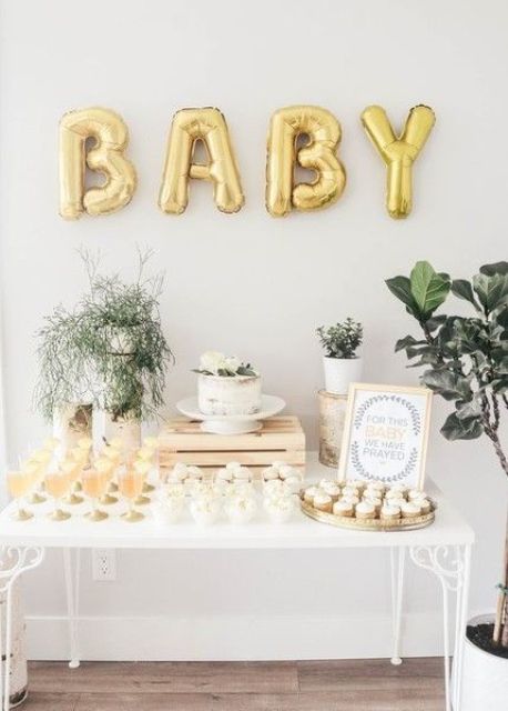 a neutral and gold baby shower dessert table with gold balloons, greenery arrangements and neutral desserts