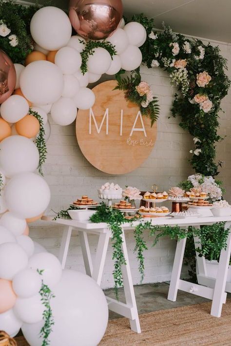 a neutral dessert table with a balloon garland with greenery and blooms, a name sign, a trestle table and greenery garlands