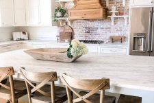 a neutral farmhouse kitchen with white cabinets, a red brick backsplash and some metal lanterns over the island