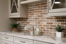 a small farmhouse kitchen with beige cabinets with a red brick backsplash and marble countertops