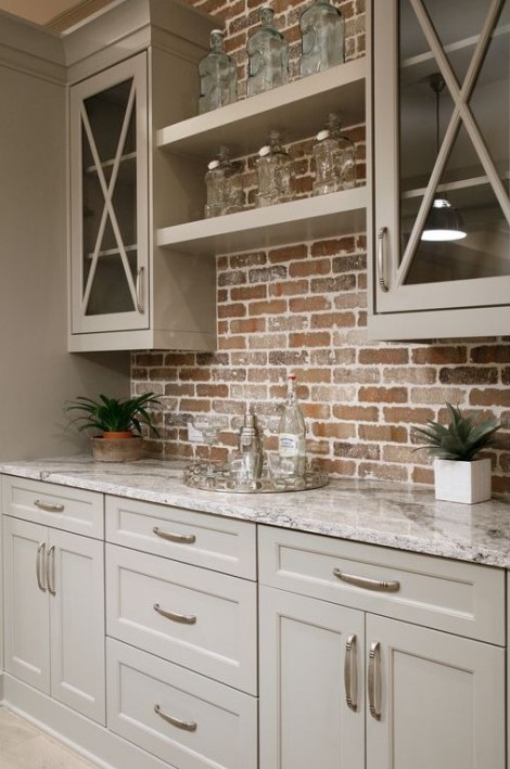 a small farmhouse kitchen with beige cabinets with a red brick backsplash and marble countertops