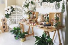 a tropical dessert table with a tropical greenery garland, white balloons, potted plants and a trestle table with crates to serve the food