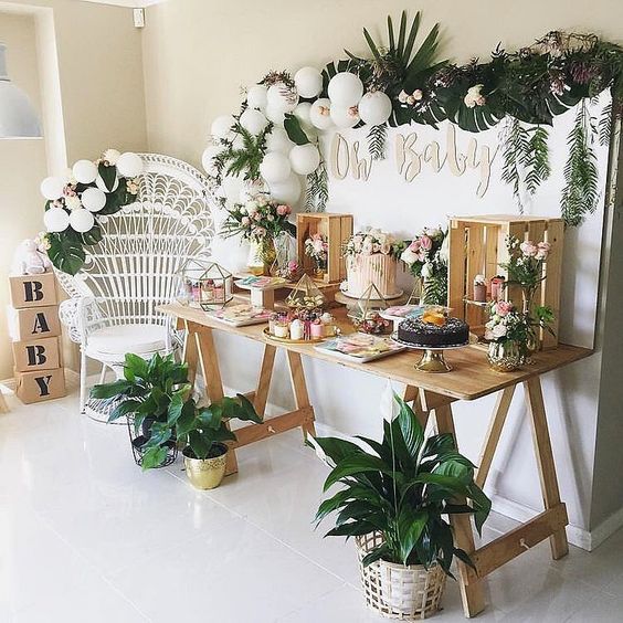 a tropical dessert table with a tropical greenery garland, white balloons, potted plants and a trestle table with crates to serve the food