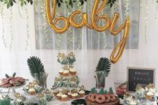 a tropical dessert table with gold calligraphy balloons, tropical greenery, a sign and lots of delicious sweets