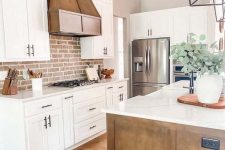 a welcoming white farmhouse kitchen with shaker style cabinets, a metal hood, dark-stained wooden beams, a red brick backsplash