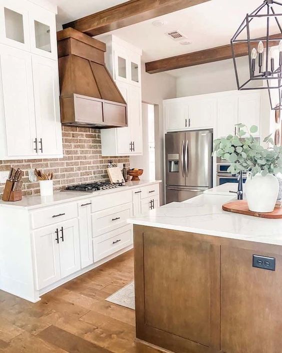 a welcoming white farmhouse kitchen with shaker style cabinets, a metal hood, dark stained wooden beams, a red brick backsplash