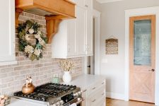 a white farmhouse kitchen with white stone countertops, a whitewashed red brick backsplash, a metal and wooden hood and some rustic decor