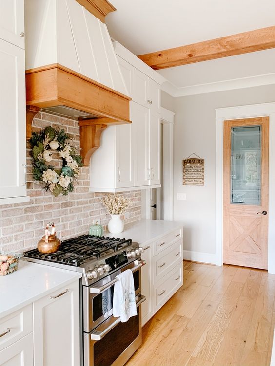a white farmhouse kitchen with white stone countertops, a whitewashed red brick backsplash, a metal and wooden hood and some rustic decor
