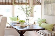 a farmhouse green and brown breakfast nook with a corner seating, a dark-stained table and various pillows, woven shades and green branches