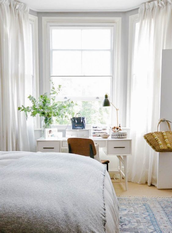 a Scandinavian grey bedroom with a bed and a printed rug, a white desk and vanity at the window, a stained chair and some greenery