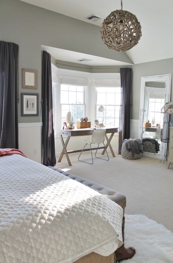 a beautiful grey bedroom with a large bed, a grey bench, a bay window and a trestle desk there, a white chair, a driftwood lamp