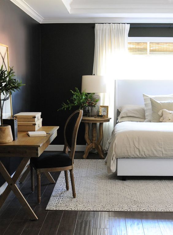 a black farmhouse bedroom with a white bed and neutral bedding, a stainted trestle desk, a black chair, greenery to refresh the room