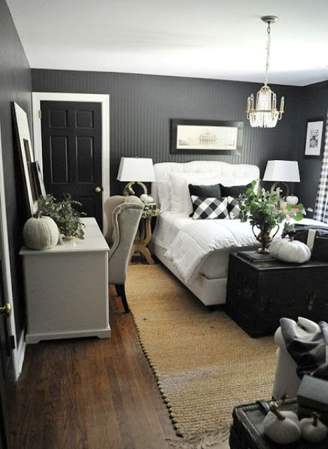 a black farmohuse bedroom with slat walls, a white bed, a black chest, a white desk and an armchair, greenery and a pendant lamp