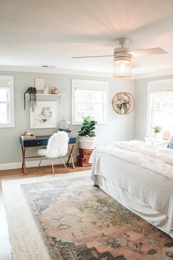a boho mid-century modern bedroom with a large bed and a printed rug, a navy desk, a faux fur chair, some plants and lamps