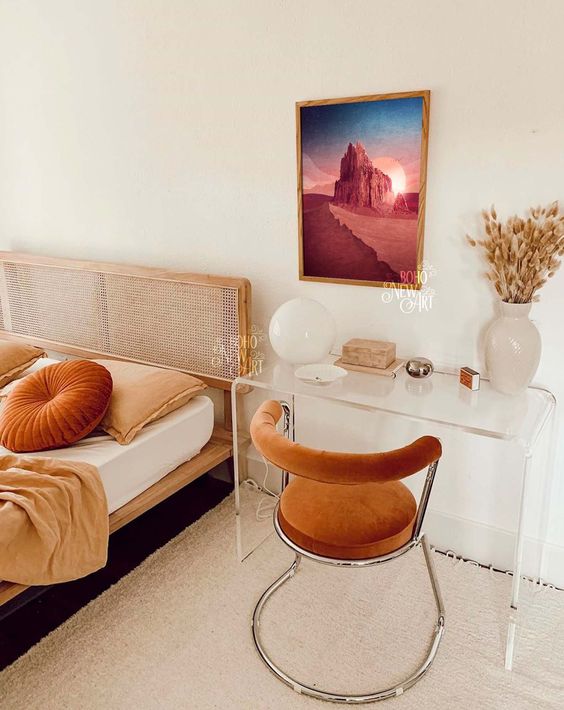 a catchy boho bedroom with a wod and cane bed, rust and peachy bedding, an acrylic desk, a rust-colored curved chair and a bold artwork