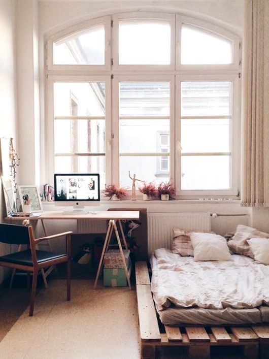 a cool bedroom with a low pallet bed with neutral bedding, a trestle desk and a black chair, some decor and boxes for storage