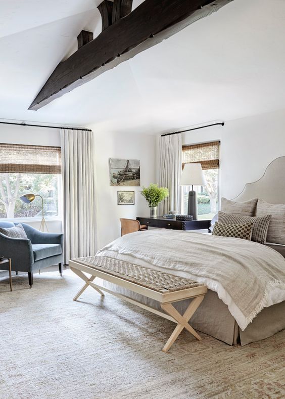 a cozy neutral bedroom with a bed and neutral bedding, a woven bench, a black desk, a leather chair, a blue chair and some neutral curtains