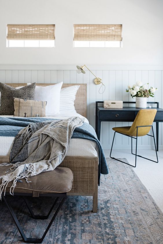 a farmhouse bedroom with white shiplap walls, a cane bed with blue and white bedding, a black desk, a leather chair and a leather bench