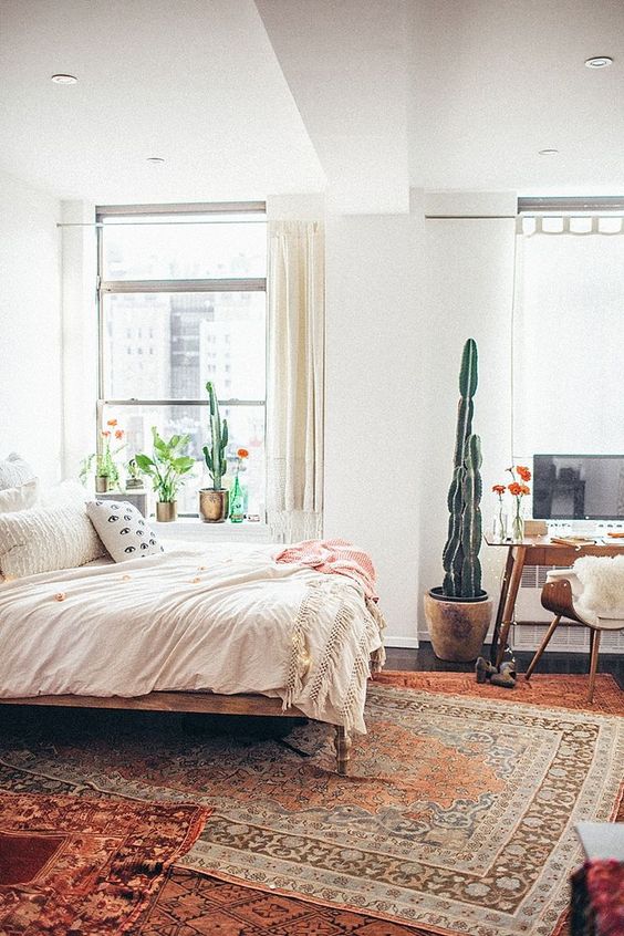 a lovely mid-century modern bedroom with a large bed and neutral bedding, layered rugs, a stained desk, a chair and some potted plants