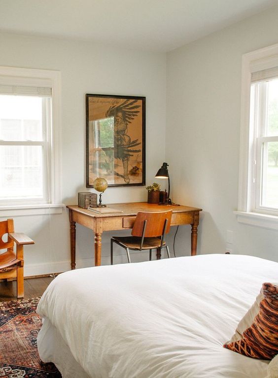 a mid-century modern bedroom with a bed with neutral bedding, a small vintage desk and stained chairs, a printed rug and artwork