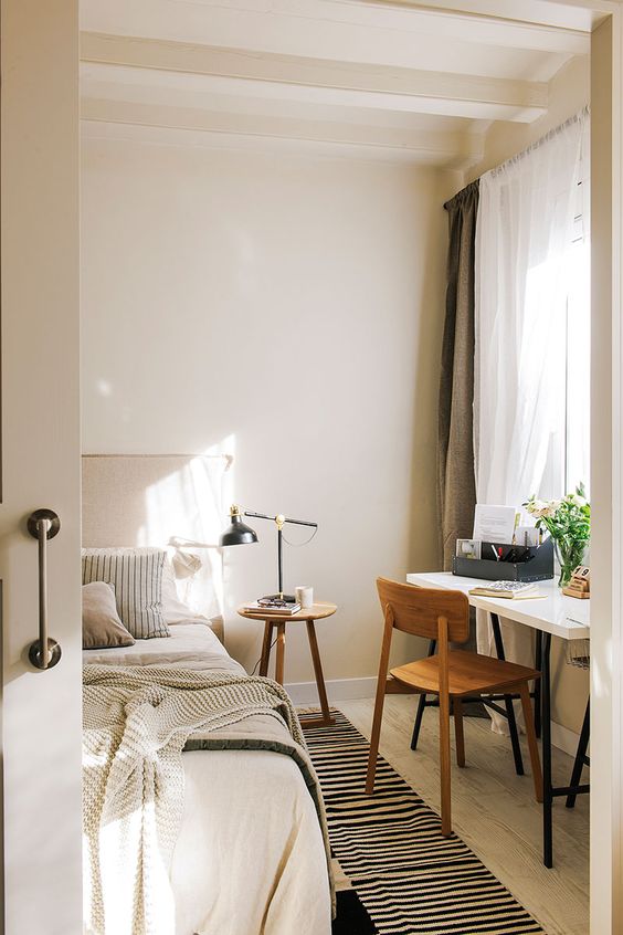 a neutral bedroom with a neutral bed and bedding, a striped rug, a desk and a chair, a wooden stool and some neutral curtains