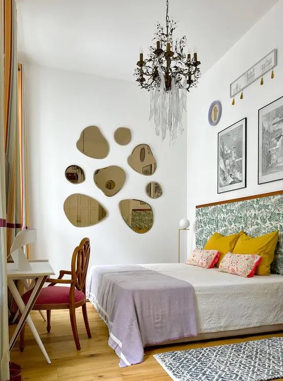a whimsical bedroom with a bed with a printed headboard, colorful and printed bedding, some art, a gallery wall of mirrors, a desk and a chair