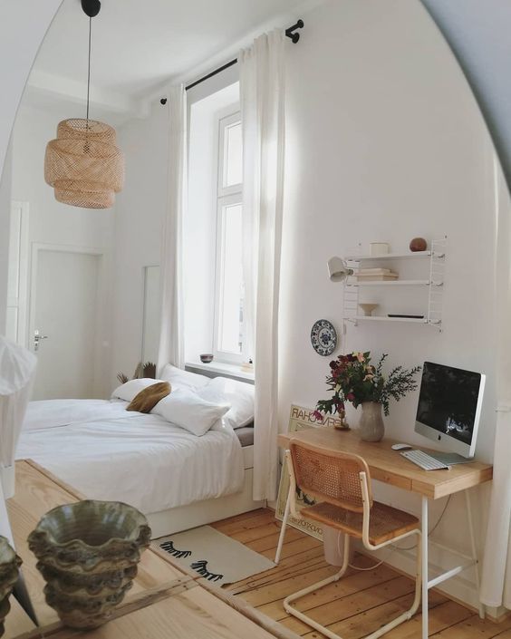an airy and light bedroom with a large bed and neutral bedding, a stained desk and a cane chair, open shelves and a pendant lamp